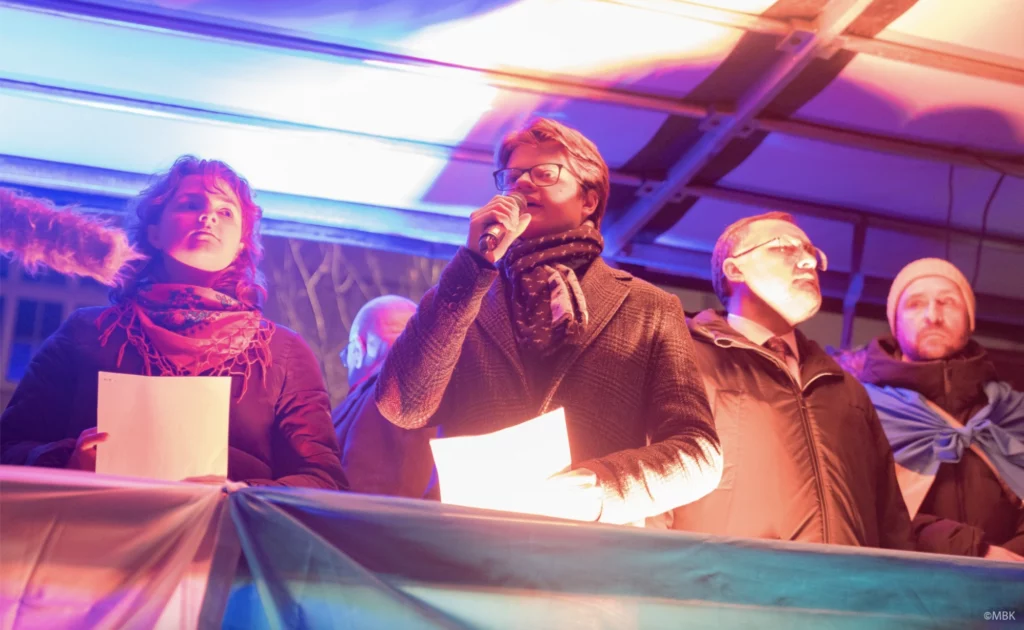 Osoby przemawiają na scenie nocnej demonstracji w kolorowym świetle, jedna z kartką w dłoni – rocznicowa demonstracja solidarności z Ukrainą
