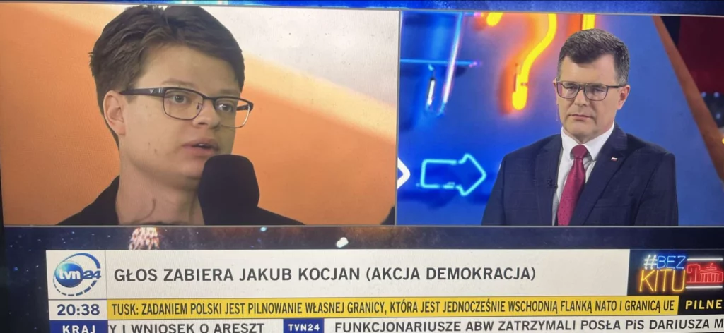 Jakub Kocjan w studiu TVN24 „Bez Kitu” na podzielonym ekranie z politykiem w garniturze, pasek „Głos zabiera Jakub Kocjan (Akcja Demokracja)”