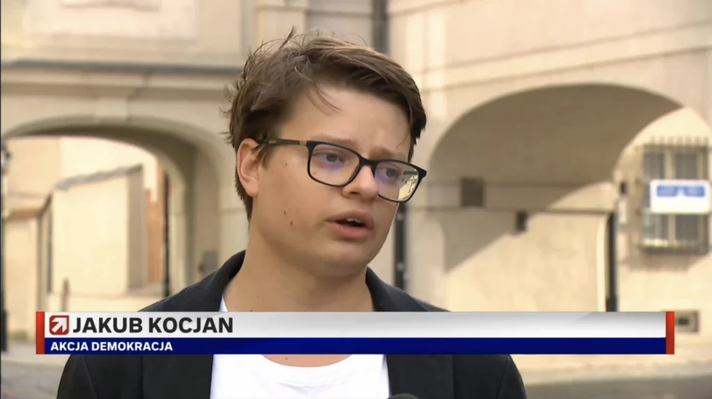 Jakub Kocjan z Akcji Demokracji w okularach wypowiada się przed kamerą na tle kamienic – kadr z „Wydarzeń” Polsatu