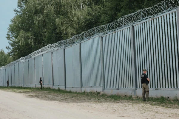 Zapora na granicy polsko-białoruskiej, metalowe przęsła z drutem kolczastym wzdłuż leśnej drogi, przy niej funkcjonariusze Straży Granicznej